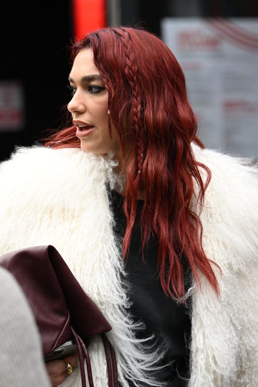 Dua Lipa wears a fuzzy cream coat, jeans, and maroon accessories.