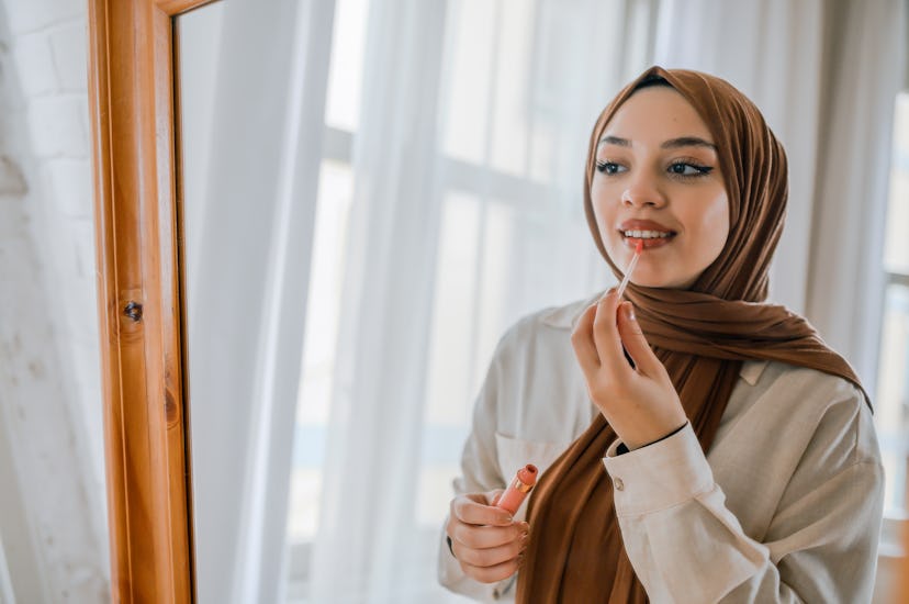 Young muslim woman wearing hijab applying lipstick in front of mirror in room