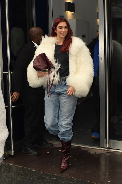Dua Lipa wears a fuzzy cream coat, jeans, and maroon accessories. 