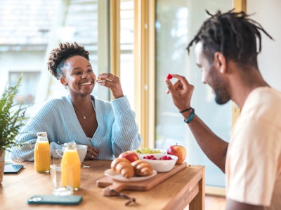 Indoor shot of a black couple sitting at a kitchen table, surrounded by a healthy breakfast spread i...
