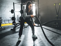 Focused young woman swinging battle ropes in alternating waves in gym