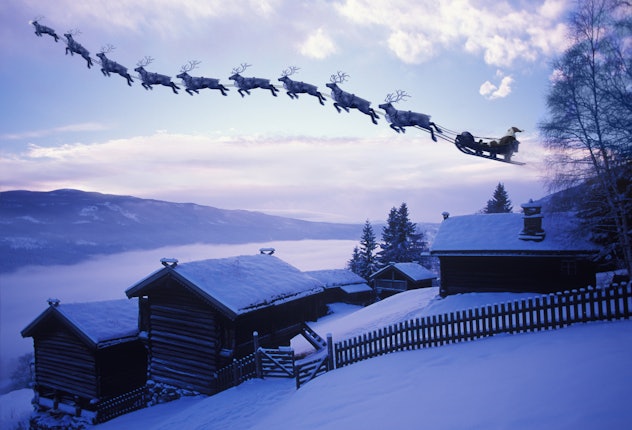 Santa Clause and his reindeer flying above an old farm covered with pristine snow