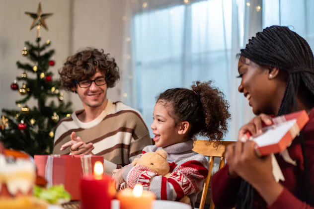 Multi-ethnic family exchanging presents during Christmas party at home. Attractive kid daughter rece...