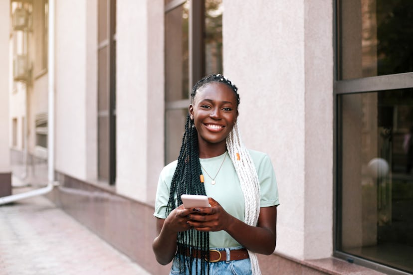 Young beautiful black braided woman using smart phone outdoors at summer at the city street. She is ...