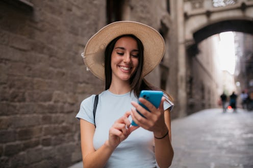 Cheerful young female on holiday using cell phone while walking in city. Woman traveler using a mobi...