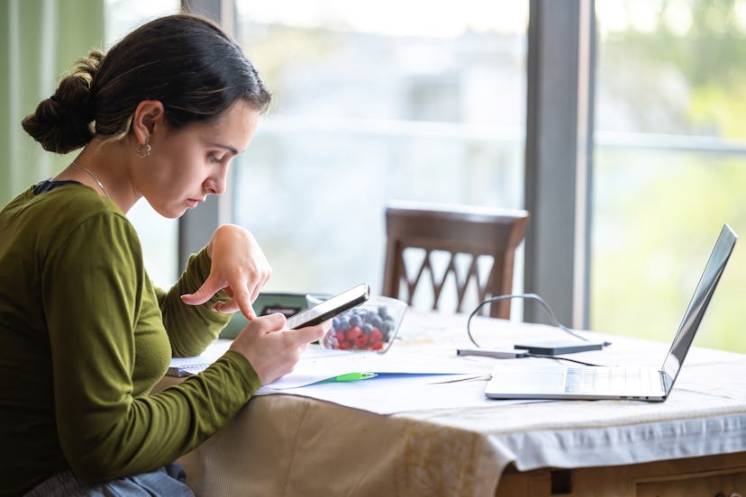 portrait of young woman sitting at table with laptop at home and typing on mobile