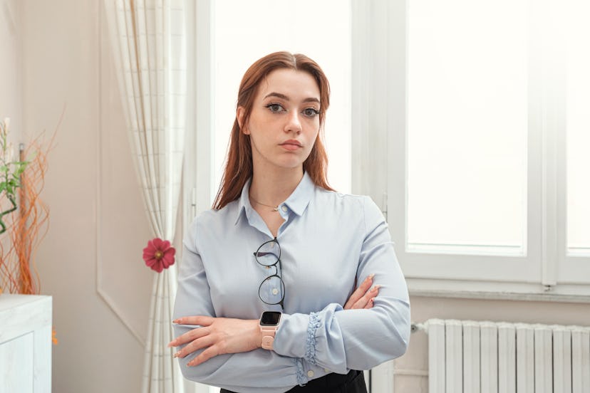 Portrait of serious redheaded woman standing in front of the window, arms crossed