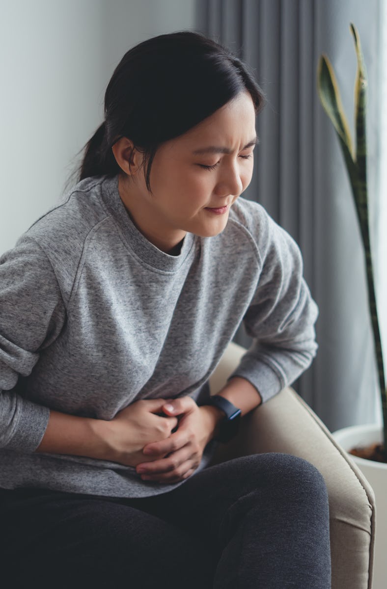 Asian woman feeling stomachache and touching her belly sitting on armchair in living room at home.