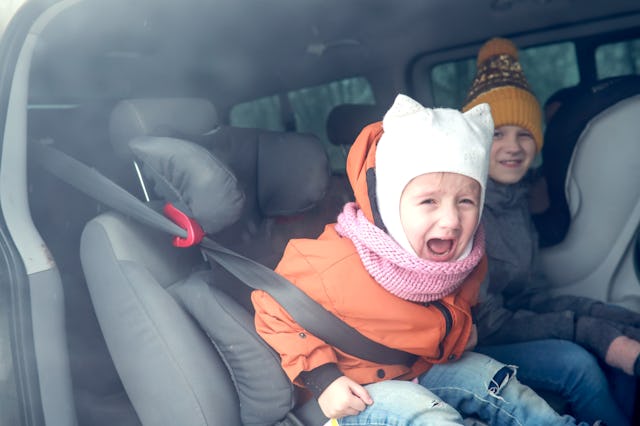 Sibling children quarrel and cry in back seat of car, older sister teases toddler in winter jacket a...