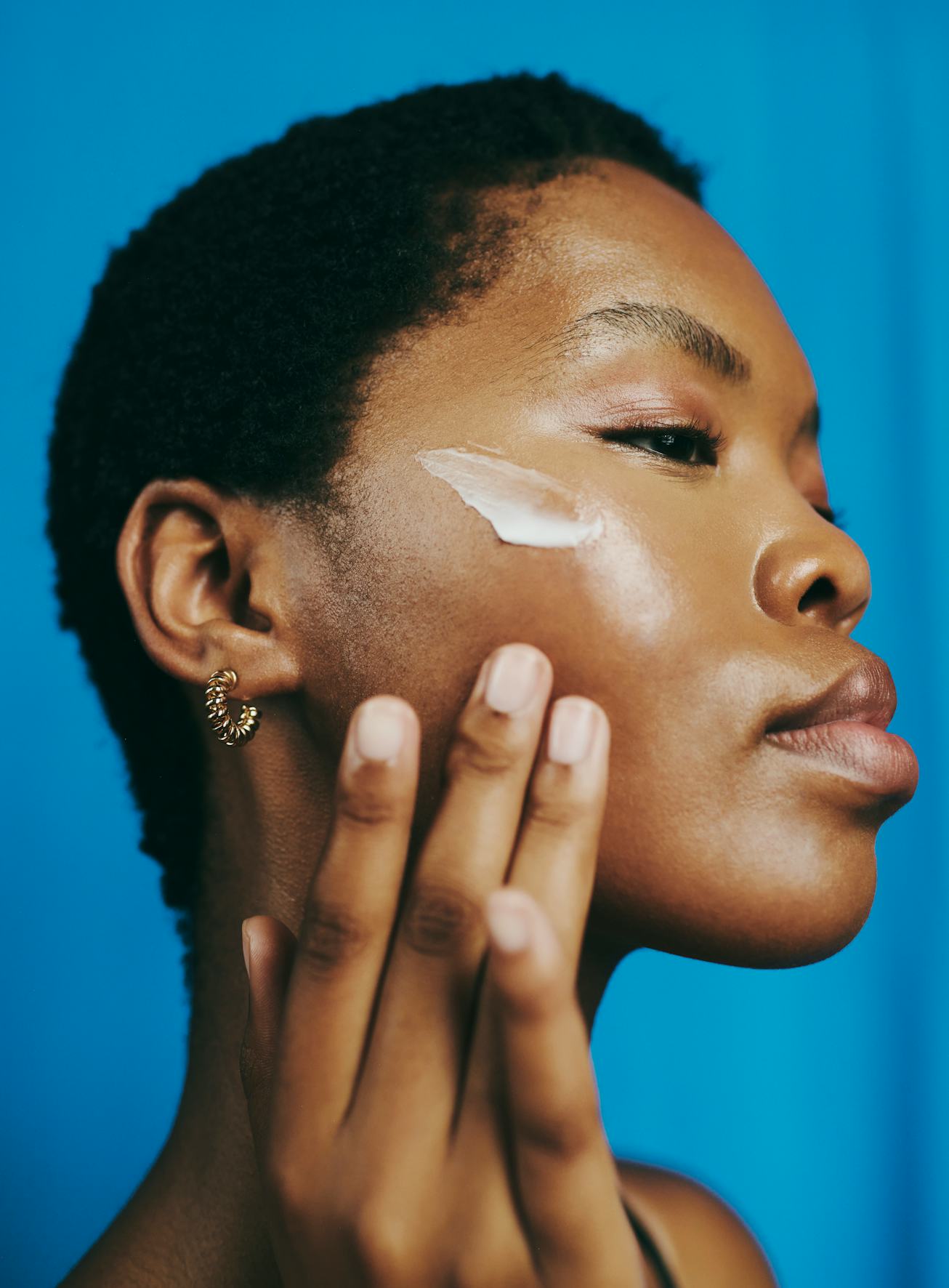 Beautiful headshot of a young black woman, looking down and thinking as she applies moisturizer to ...