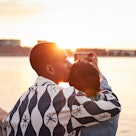 Young man kissing woman while photographing river on mobile phone during sunset