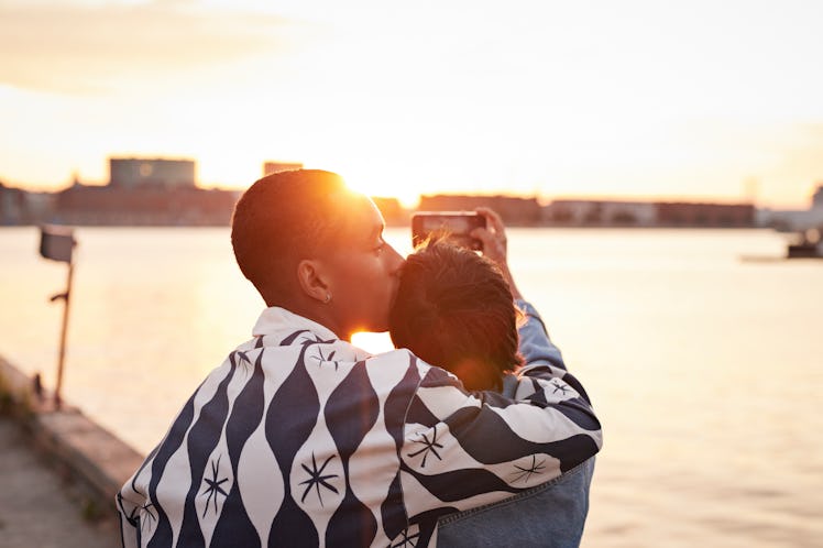 Young man kissing woman while photographing river on mobile phone during sunset