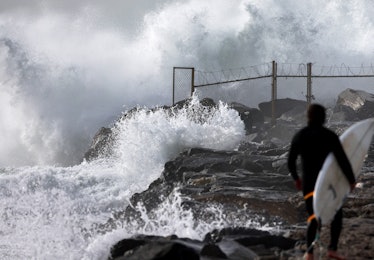 A surfer walks along a jetty on his way to enter the water as large waves break near the beach on De...