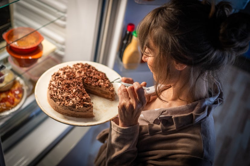 Mature woman eating cake while standing near open refrigerator at home.