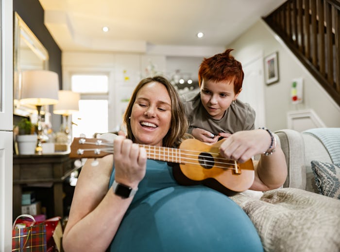 Expecting mother and her daughter relaxing at home. Mother is playing ukulele while daughter is giv...