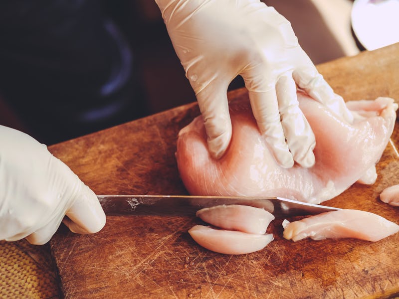 High angle shot of a person in gloves slicing raw chicken fillet on old wooden cutting board.