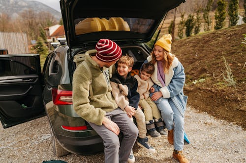 Photo of a smiling, cheerful family sitting in the trunk of their car, all packed up and ready for a...