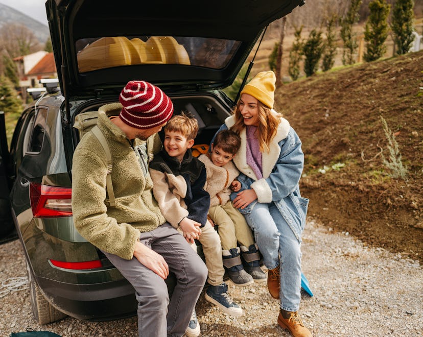 Photo of a smiling, cheerful family sitting in the trunk of their car, all packed up and ready for a...