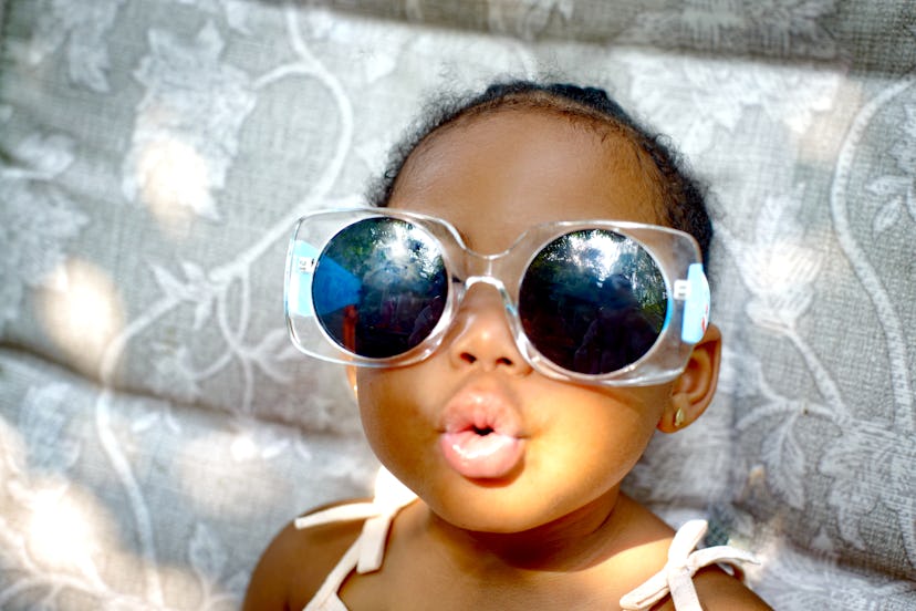 Baby girl in sunglasses blows kiss to camera, in Romper’s baby name trend predictions for 2024.