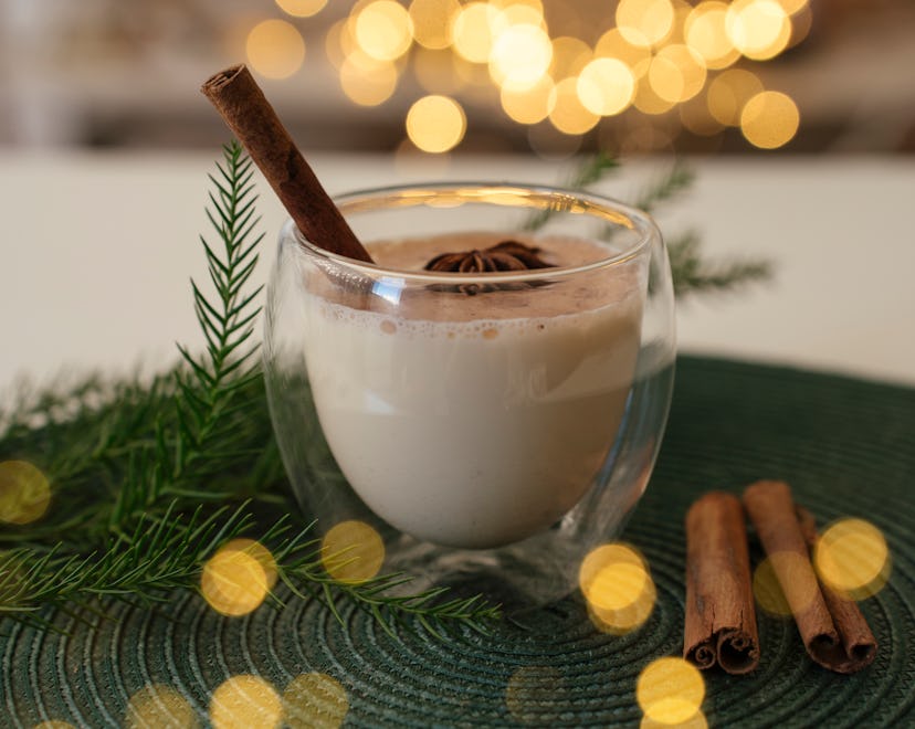 Traditional Christmas dring Eggnog with cinnamon in glass on white table. Festive setting, green pin...