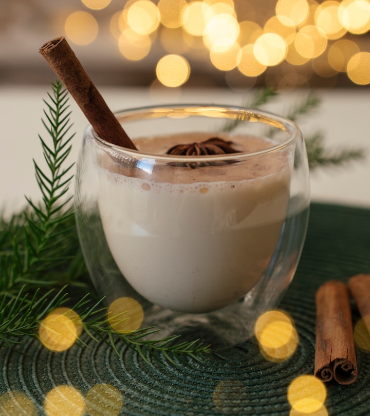 Traditional Christmas dring Eggnog with cinnamon in glass on white table. Festive setting, green pin...