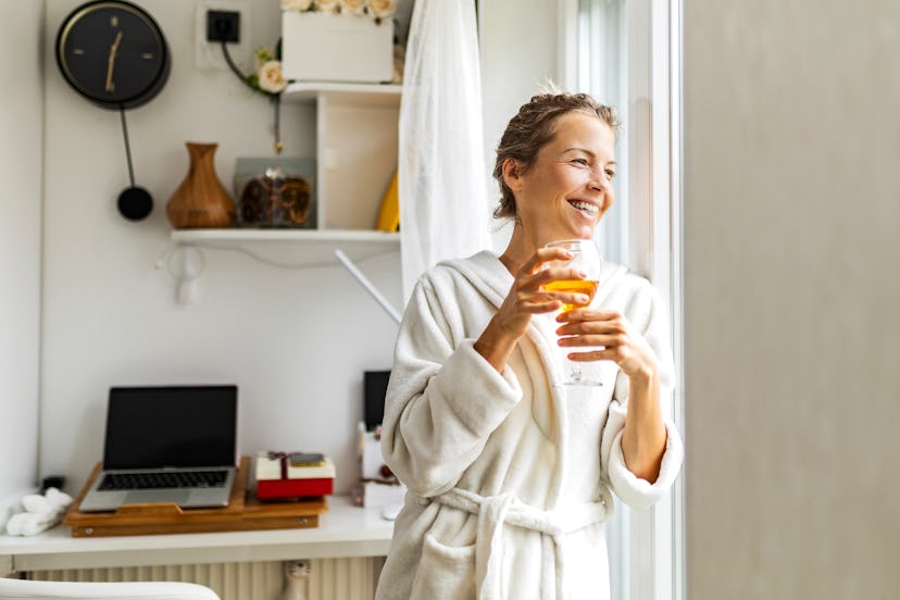 Attractive young woman holding wine glass looking through the window wearing bathrobe