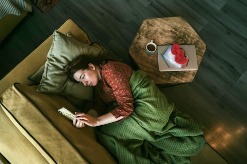 Portrait of a red-haired woman covered with a green blanket lying down on the yellow sofa with mobil...