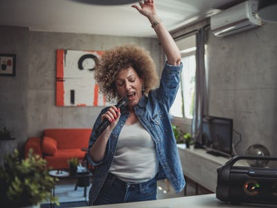 One woman, a cute middle-aged woman with curly hair is having fun at home singing karaoke, holding a...