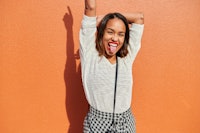 young woman sticks out her tongue as she poses for a photo against an orange wall, and considers whi...