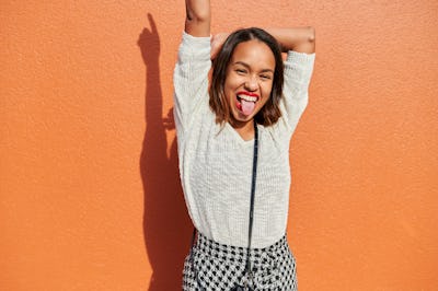 young woman sticks out her tongue as she poses for a photo against an orange wall, and considers whi...