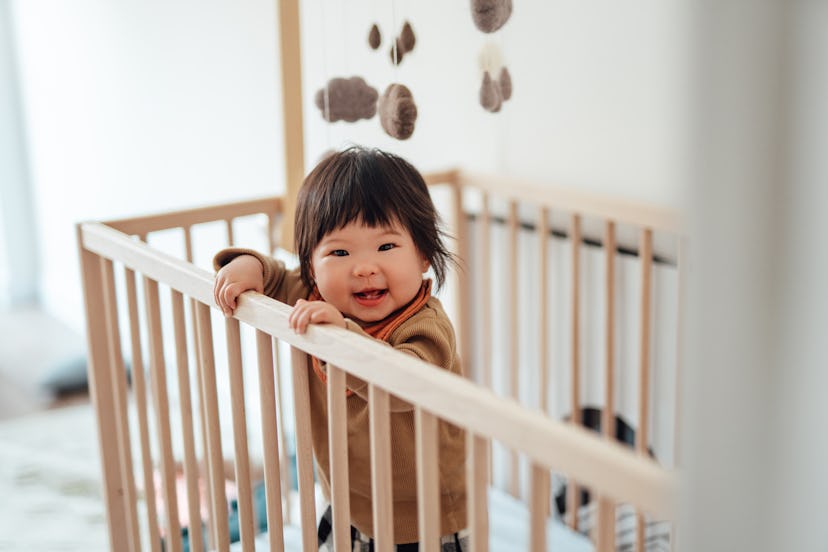Headshot of a cheerful baby girl learning to stand in the crib, in an article about the 8 month slee...