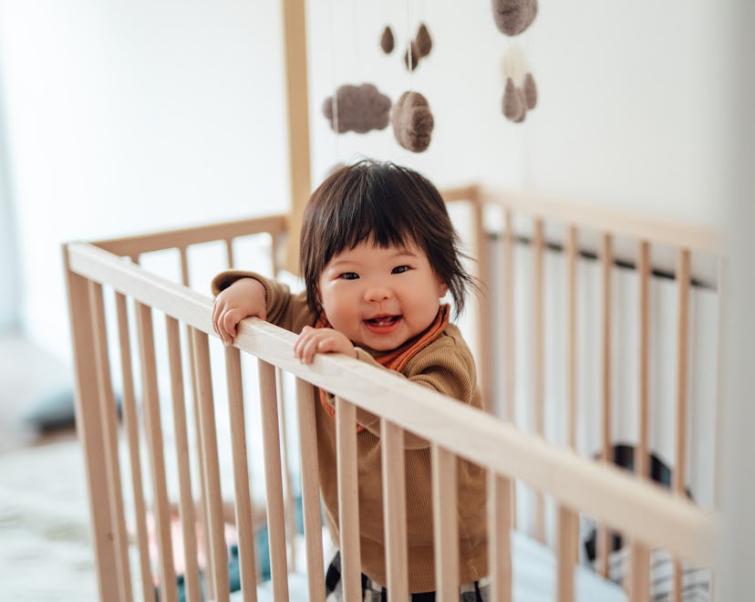 Headshot of a cheerful baby girl learning to stand in the crib, in an article about the 8 month slee...