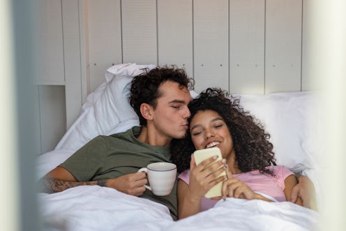 Couple sitting in bed in the morning together drinking to drinks while on a staycation. They are sta...