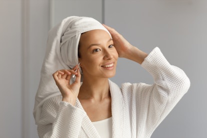 Pretty woman in white robe and with towel on her head cleans her ears with cotton stick. Cleansing a...
