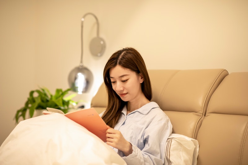 Young woman lying on the bed and reading a book at night. The room is dark and the light on the nigh...