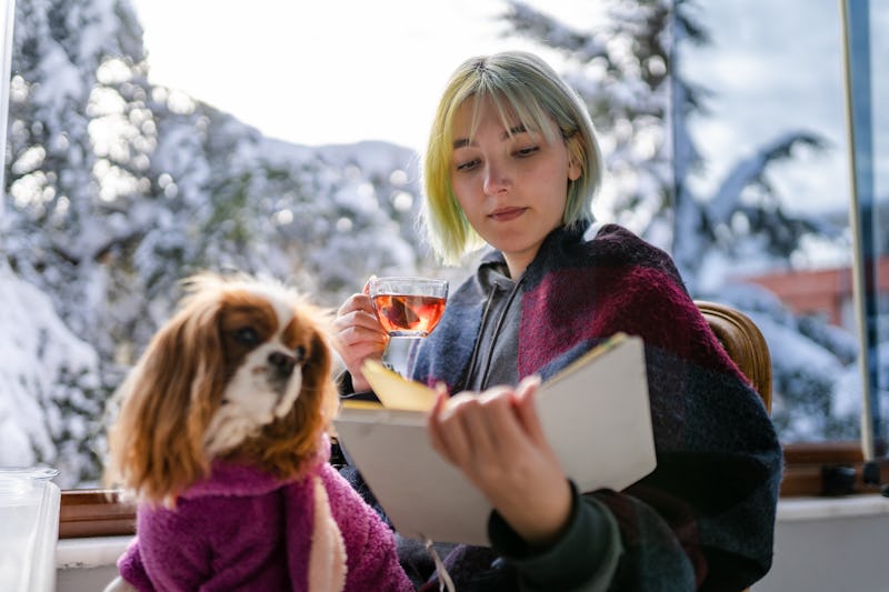 Young girl sitting on the balcony of her house with her dog while it is snowing, reading a book and ...