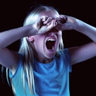 A little blonde girl is seen against a black background, her hands covering her eyes and her mouth w...