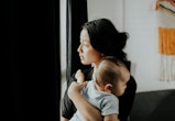 Young woman carrying baby looking out window, in a story about postpartum psychosis.