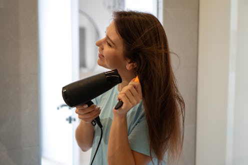 Young woman dries her long brown hair with a hair dryer looking to the mirror. Attractive female pro...