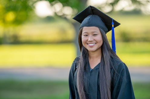 A young female graduate stands out side as she poses for a portrait. She is wearing a gown and cap ...