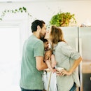 A couple in their kitchen holds their baby between them and kisses.