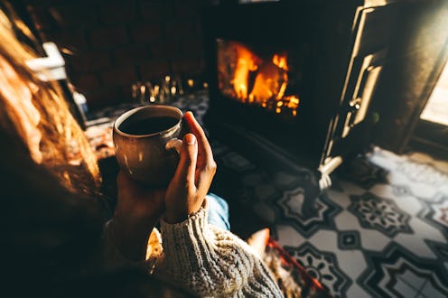 Woman relaxing in a knitted wool sweater, sitting in front of the fireplace, embraces a mug with her...