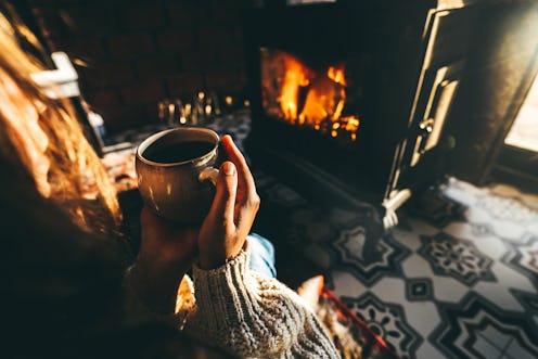 Woman relaxing in a knitted wool sweater, sitting in front of the fireplace, embraces a mug with her...