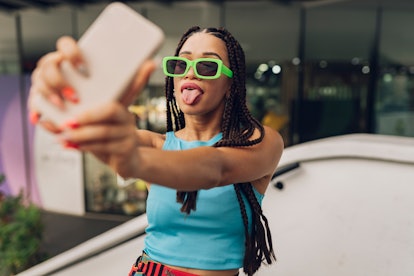 a young woman wearing green glasses sticks out her tongue while taking a selfie, as she considers wh...