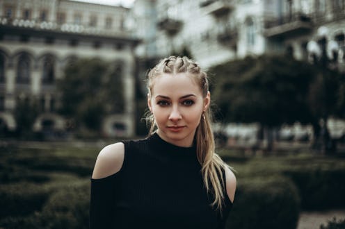 Beautiful young woman in black clothing standing against blurred trees and vintage buildings.
