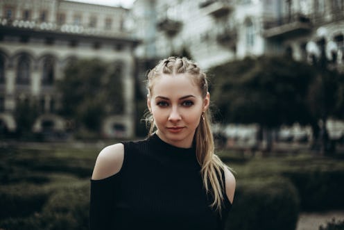 Beautiful young woman in black clothing standing against blurred trees and vintage buildings.