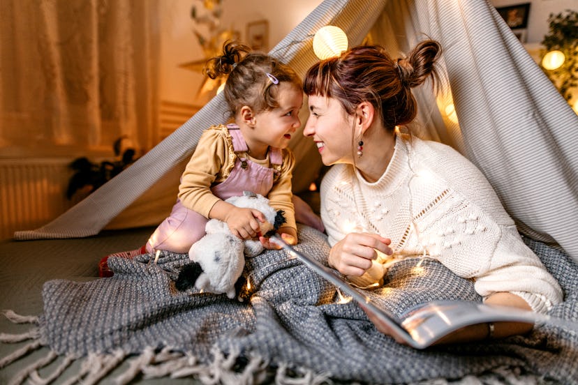 Mother and daughter are having fun reading a book under the illuminated tent in the bedroom. is mela...