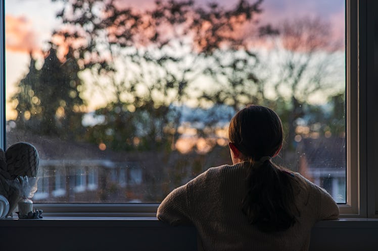 A young kid looks out of their dark bedroom window.