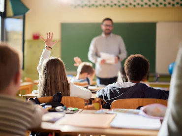 Rear view of schoolgirl raising her hand to answer the teacher's question on a class at elementary s...