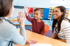 A cheerful teacher gives a little boy a high five during a parent-teacher conference. The boy's mom ...